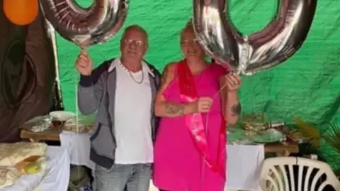 Michelle Maher A man in a white t-shirt and grey jacket and a woman in a pink dress smile holding large birthday balloons.