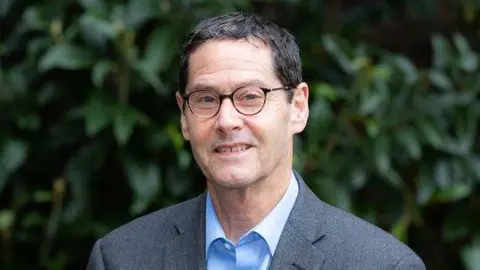 CUHNHSFT David Rowitch is in the middle of the frame. He is looking at the camera and has short hair and is wearing glasses, a blue shirt and grey suit jacket. He is standing in front of a green hedge. 