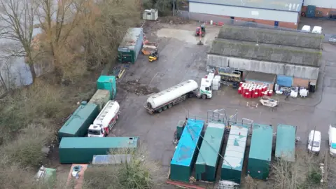 Lee Cripps A drone photo showing oil tankers moving around a concrete yard. There's some trees and the edge of a lake directly next to it.