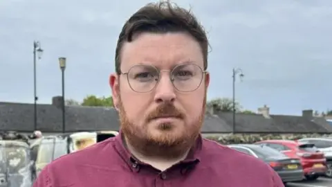 Richard Brown Stewart has short brown hair and a beard. He is wearing a purple shirt. He has circular glasses and is standing in a car park. 