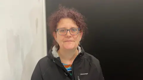 A woman with dyed purple curly hair, glasses and a black jacket looks directly into the camera.