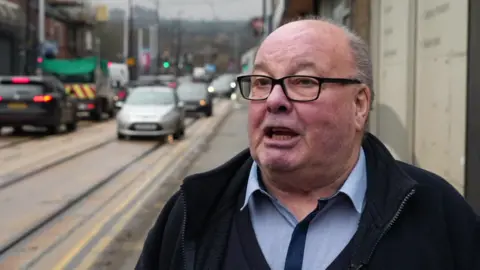 A man in his seventies stands against the backdrop of a busy road. A queue of traffic as well as some tram tracks can be seen over his right shoulder.