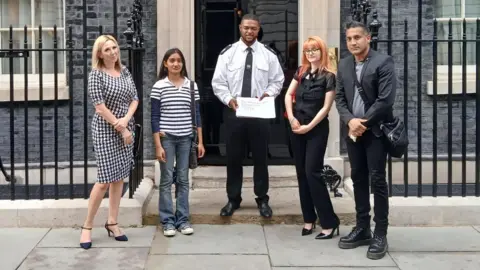 Wilson Chowdhry Five people stand in front of the door to 10 Downing Street. A police officer stands in the middle holding a piece of paper. 