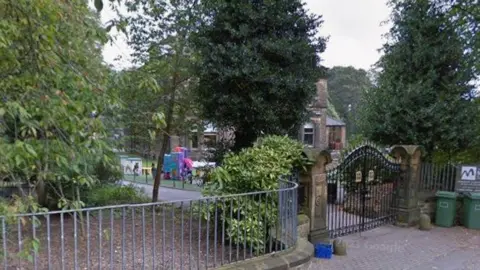 A gated property, pictured from behind large iron gates. Lots of tall trees are within the grounds of the property. Some children's play equipment can be seen within the grounds.