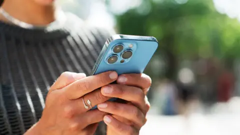 Getty Images A close up of a person's hands holding a blue iphone, which we can only see the back of.