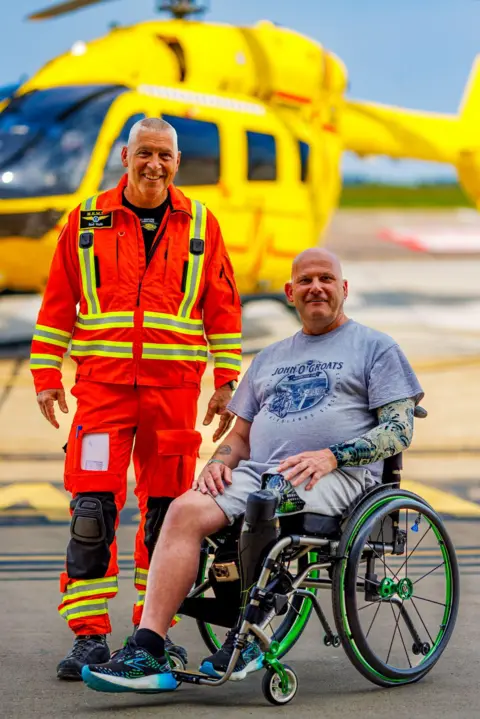 EAAA A man in a wheelchair and a man wearing a paramedic's uniform face the camera with a yellow air ambulance helicopter in the background behind them.