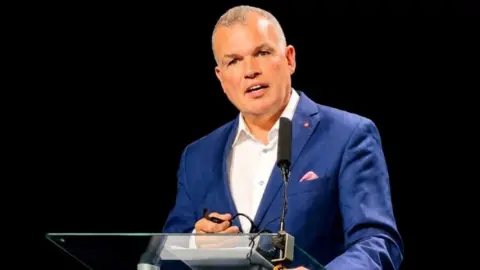 Doncaster Chamber A man is wearing a bright blue suit and a white open neck shirt and is stood speaking at a podium with a black backdrop