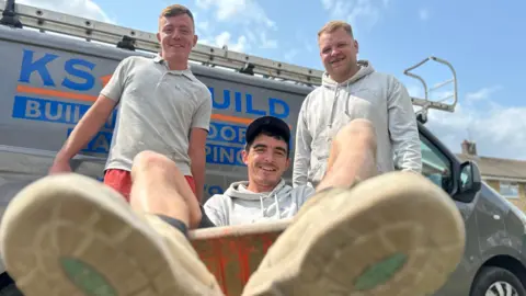 Three men, all wearing grey and smiling with one man sat in a wheelbarrow and the other two holding either handle