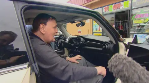 Jim Torbett sitting in a car with the drivers door open. A microphone is being pointed at him.