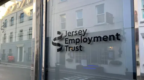 The glass, exterior door to an office in St Helier. On the glass are the words 'Jersey Employment Trust' in black writing, next to a black logo. The door frame is shiny chrome. Another, four storey building is visible in the reflection in the glass.