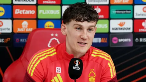 PA Media A mid-twenties footballer wearing a red and yellow tracksuit speaking into a microphone.