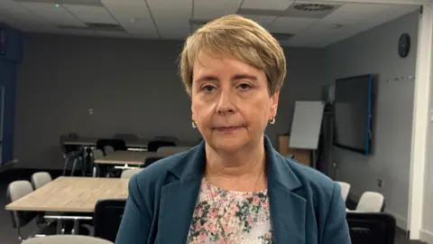 Gill Findley stands inside an office room which has a collection of empty tables and chairs. She is wearing a flowery top and navy suit jacket. She has short brown hair.