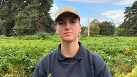 There is a woman with a beige cap on. She is wearing a dark blue jumper and a black polo-shirt. She has a nose ring on her right nostril and her hair is tied behind her back. Behind her is green shrubbery, big dark green trees and a bare white tree.