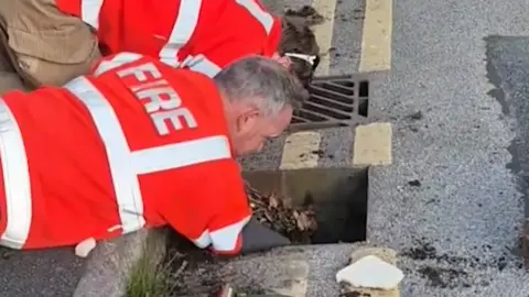 A man wearing a bright orange high-vis top with "fire" on his back reaches into an open drain. A woman in the same clothing assists next to him.