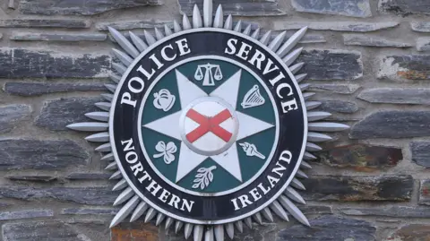 Police Service Northern Ireland badge against a brick wall.

The badge is black on the outside, green in the middle with justice symbols and then the inner has a star with a red St George's cross.

 

 