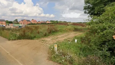 Google The site of 35 proposed houses on Station Road, Digby. There is a farm track leading into a field in the foreground, and behind the field are new-build houses and construction items.