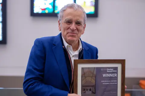 Stuart Hay John McAslan in a blue jacket stands in front of the Burrell's stained glass holding the Andrew Doolan award - a framed plaque with "winner" printed on it.