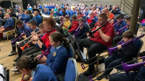 A group of children are playing a mixture brass instruments while sitting in front of music lecterns. The instruments are different colours - some purple, one green, one orange and one black.