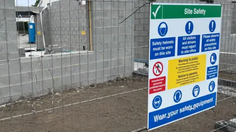 BBC Image of a construction site safety sign with a a part-built, grey-brick building behind a fence. There is a sign on the fence giving guidance on site safety.