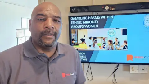 A man wearing a grey polo standing in front of a television screens, which says gambling harms within ethnic minority groups/women