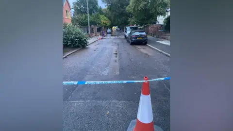 A traffic cone and police tape can be seen at one side of a road, with some water seen on the road and an emergency services vehicle at the other end of the road.