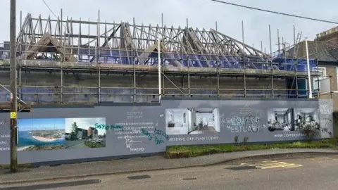An incomplete building with no roof is surrounded by scaffolding. Advertising hoardings offering modern flats are covered in Graffiti .