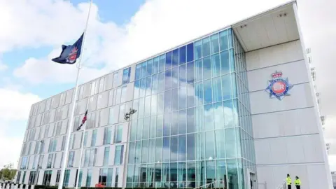 A wide shot of the Greater Manchester Police building. It is a covered with windows and has a grey exterior. The force's badge is also on the side of the building.
