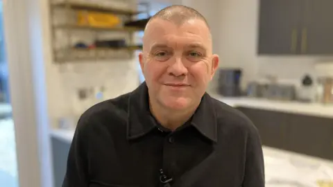 Dominic Matteo, a man in his 50s in a black shirt and shaved dark hair, stands in his kitchen. 