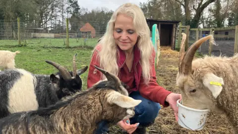 A woman with white blonde wavy hair in a red jumper is crouched down in a field. She has her arms in front of her - one is holding a white bucket. A large beige goat with horns atop its head is sticking its face into the bucket. Near her other hand, two goats are close to her - one is black and white with horns and the other is black and brown with cream coloured ears. The field beyond has green grass and is very muddy, with hay leading up to a small wooden enclosure