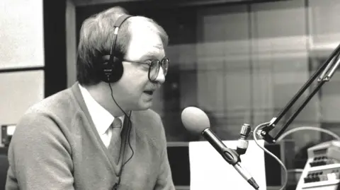 Gavin McCoy is wearing a V-neck jumper over a white shirt and tie. He is speaking into a microphone while wearing headphones.