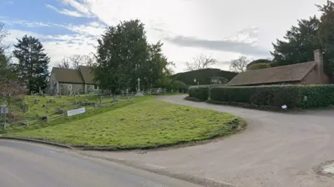 A google maps image of St. Nicholas's Church in Pyrford, near Woking, Surrey. part of the church can be seen in the image alongside an entrance and a grassy area.