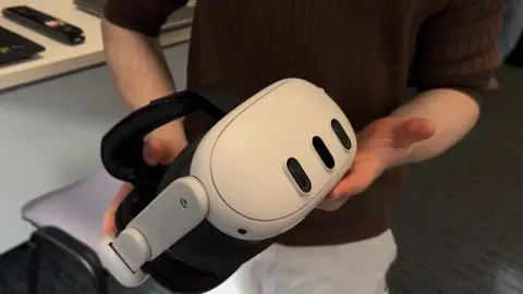 A man with a brown shirt and white trousers is holding a white VR headset.