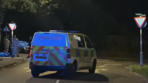 A police van is marked in the middle of the road as officers deal with the incident. Police in white overalls stand on the corner. It is night and the road signs are lit up. A police cordon can be seen in the corner.