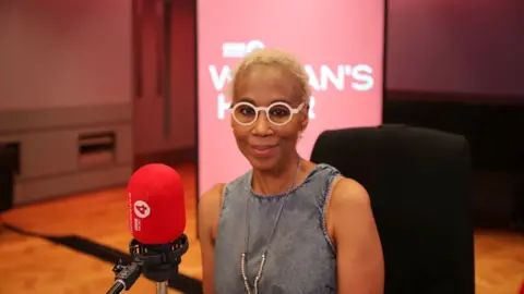 Trisha Goddard sits in front of a red Woman's Hour microphone. She is wearing large white glasses and a denim top.