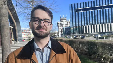 Kate Bradbrook/BBC A man with glasses and a beard, wearing a tan jacket and blue shirt, stands in front of a glass building with blue sky.