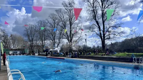Kate Bradbrook/BBC An outdoor pool, with trees at the side. Ropes divide the pool into lanes and there is bunting flying overhead. 