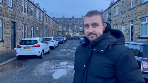 A man in a dark-coloured padded jacket with a hood standing on a terraced street with potholes.