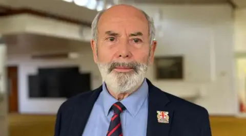 BBC A man with a grey beard in a blue suit and a stripped tie.