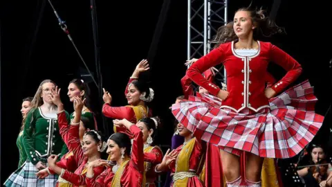 A large stage with a black background has a number of women taking part in Irish dance. The women are wearing black, red and green outfits with matching checked skirts.