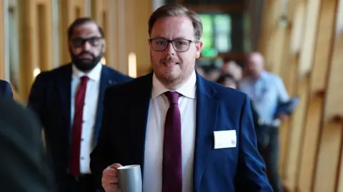PA Media Thomas Kerr, who has fair hair and glasses, walks with a mug in his right hand 
