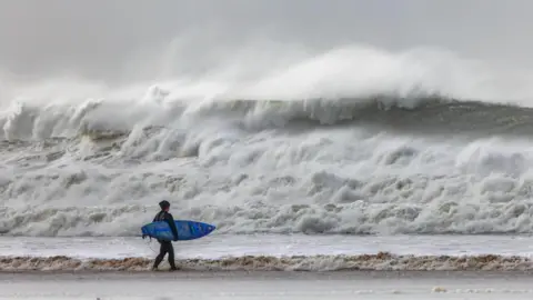 BBC Weather Watchers/FoggyLens The picture shows a lone surfer standing on the shoreline, holding a blue surfboard and looking out towards huge, churning waves. The sea is rough and wild, with towering white‑topped swells crashing in the distance. The sky looks grey and stormy.