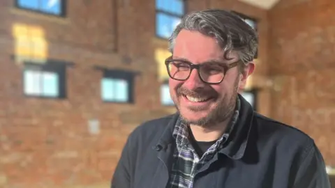 Ross Millard, smiling at the camera. He is wearing a green-and-purple checked shirt and a dark blue jacket. He has short wavy hair and glasses.