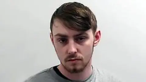 A man with short brown hair and brown facial hair looks at the camera with no expression. He wears a grey jumper.