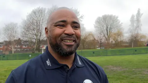 Mark Lewis-Francis stands on a grassy field. He is wearing a blue polo shirt with white writing advertising the European Athletics Championships in Birmingham in 2026. He has a shaved head and stubble. He is smiling. 
