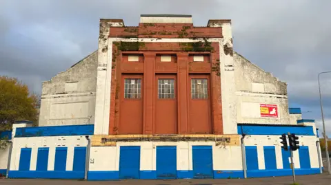 A photo of the art deco style building. The central section has three tall windows framed by reddish-brown stonework, and all ground-level entrances are covered with metal shutters. 
