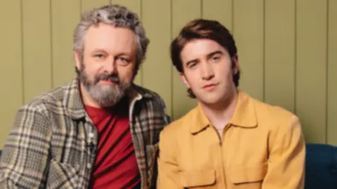 Sheen and Howells sitting next to each other on a navy sofa, looking at the camera. 