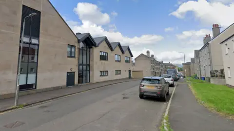 A general view of William Street in Johnstone. There are flat blocks on either side of the road. A grass patch separates the pavement from the flats on the right side. There are cars parked by the side of the road.