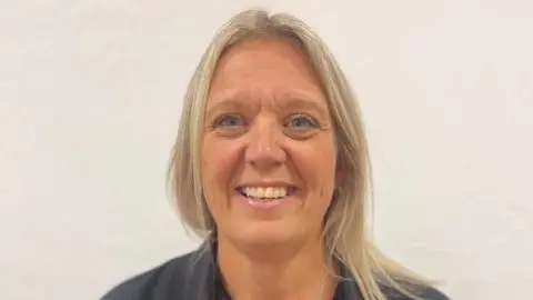 Melissa Barker-White A head and shoulders photo of Melissa Barker-White. She is smiling at the camera and is stood in front of a white wall. She has long blonde hair draped over one shoulder and is wearing a black top.