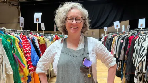 A woman with a curly grey bob wearing a white shirt and grey overalls. She is smiling at the camera as she stands in front of rails of brightly coloured clothes.