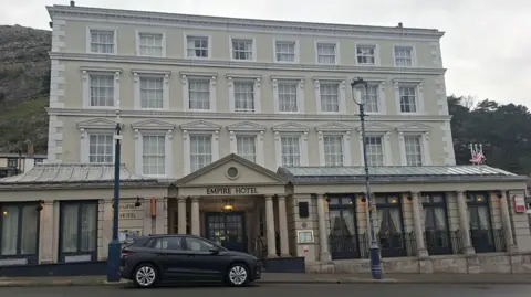 The Empire Hotel in Llandudno - it is a cream building with white windows standing three storeys tall. On the street in front of the building is a black car and the Great Orme rises behind the building in the background